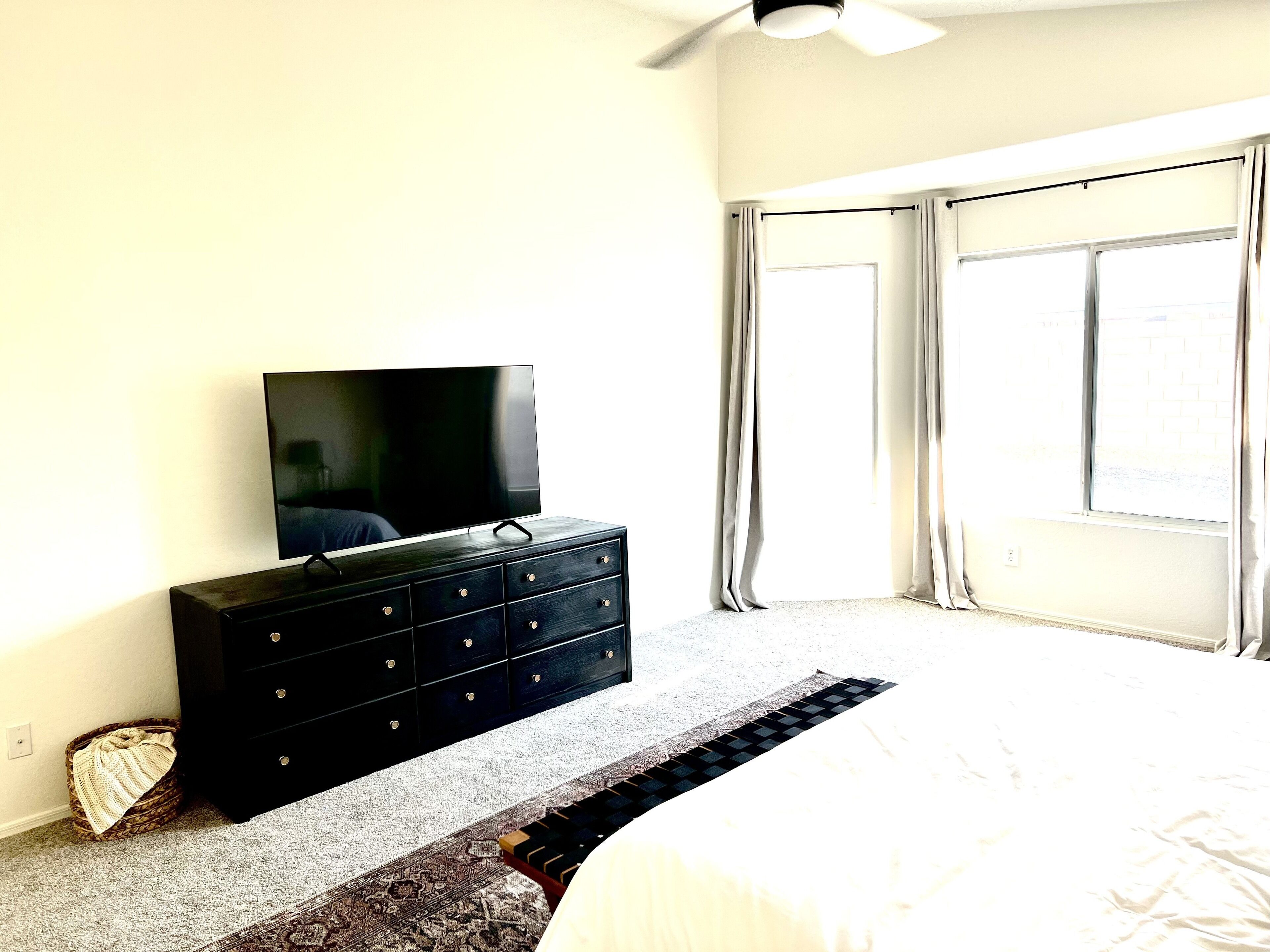 A bedroom with a bed, a dresser with a television, a window with curtains, and a ceiling fan.