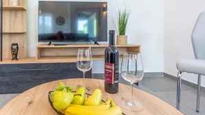 En moderne stue med et træbord, der holder en flaske vin, to glas og en kurv med frugt. Et fladskærms-tv er monteret på væggen over en træhylde med pynteting.