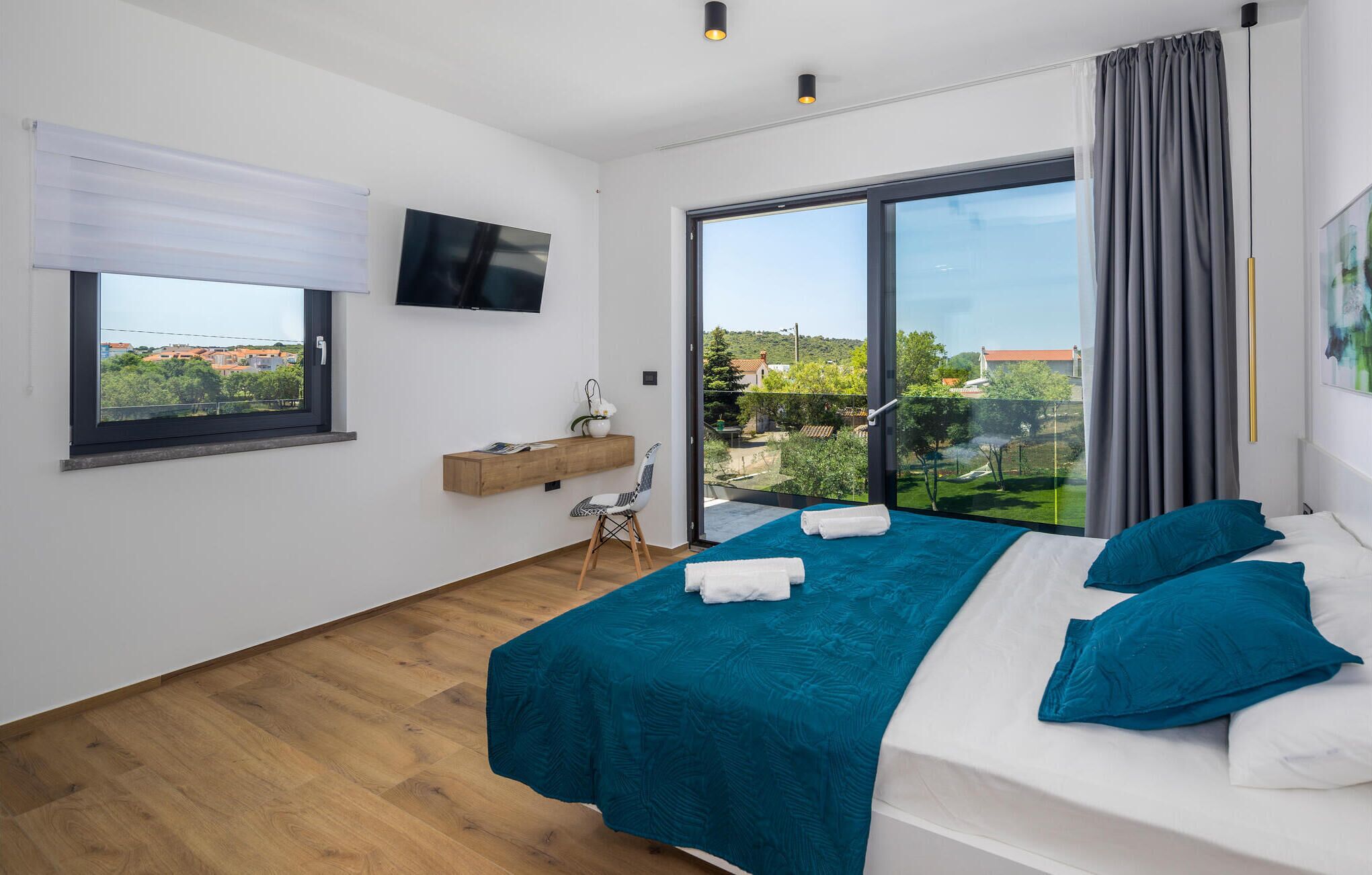 A modern bedroom with a large bed, a TV, a window with a view, and a wooden shelf.