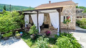 Una petita casa de pedra amb teulada de rajola, zona d'estar exterior coberta amb cortines blanques, i un jardí amb diverses plantes i flors en testos.