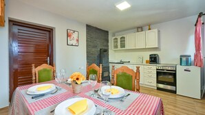 Une salle à manger avec une nappe à carreaux, des chaises en bois et un vase rempli de fleurs. On aperçoit, au fond, une cuisine avec des meubles blancs et un réfrigérateur VIVA X.