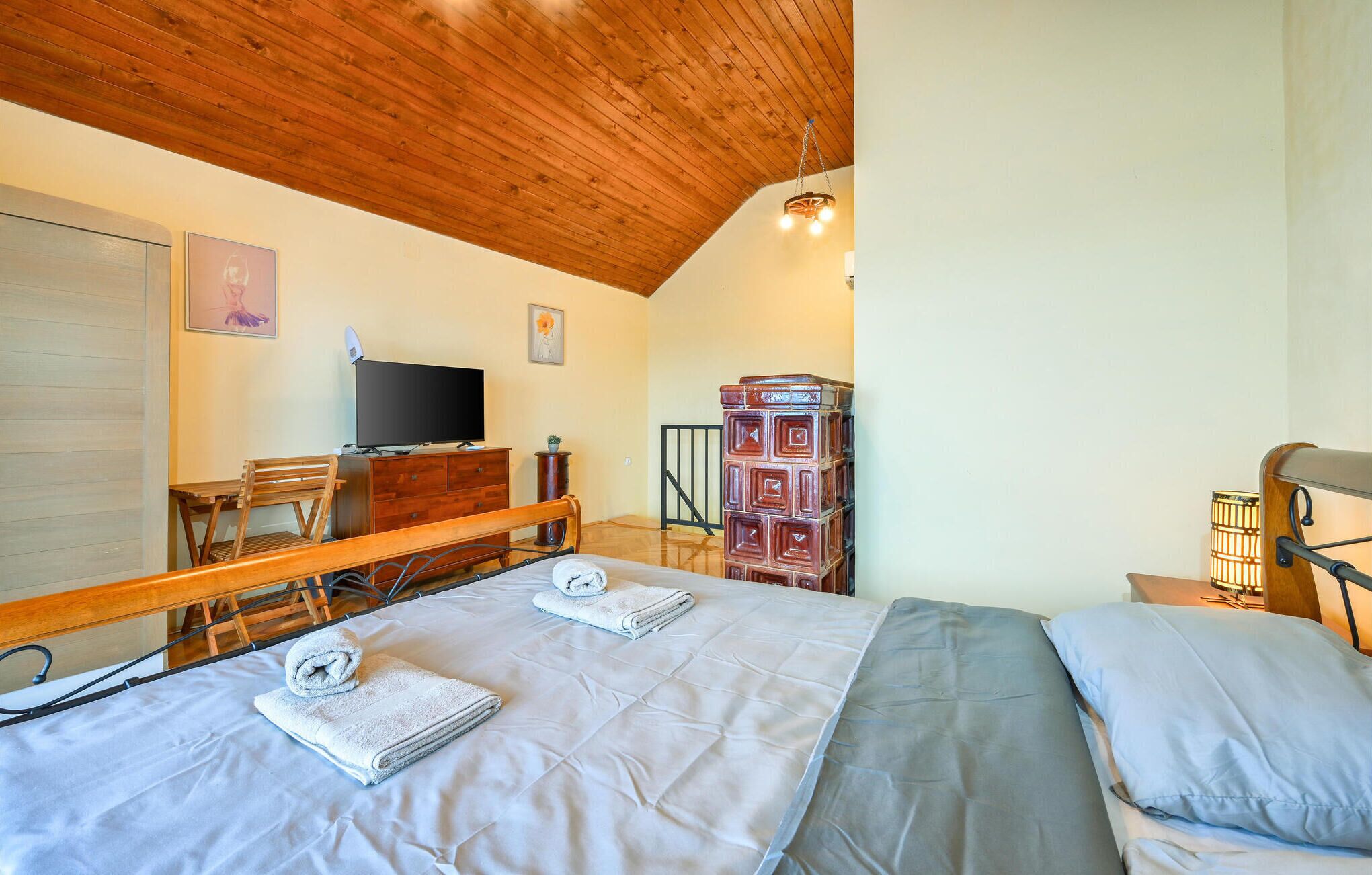 Ein Schlafzimmer mit Holzbalkendecke, einem Bett mit grauer Bettdecke, einer Holzkommode, einem Fernseher und einem kleinen Tisch mit einer Pflanze.