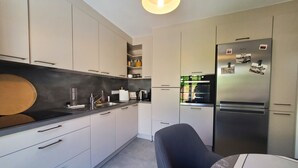 Una cocina moderna con armarios blancos, encimera negra, refrigerador de acero inoxidable y una zona de comedor con una silla.