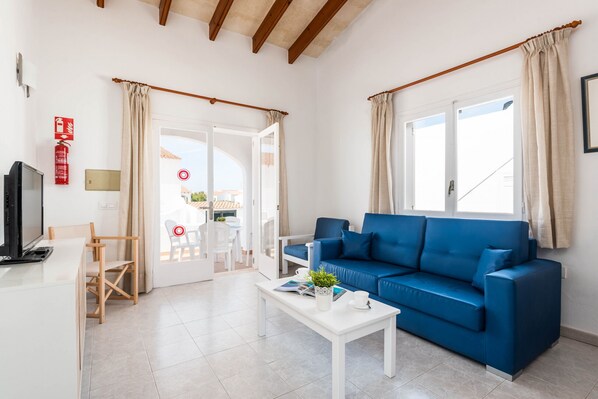 Un salon moderne avec un canapé bleu, une table basse blanche et un téléviseur. Une porte donne accès à un espace extérieur aménagé avec une table et des chaises.