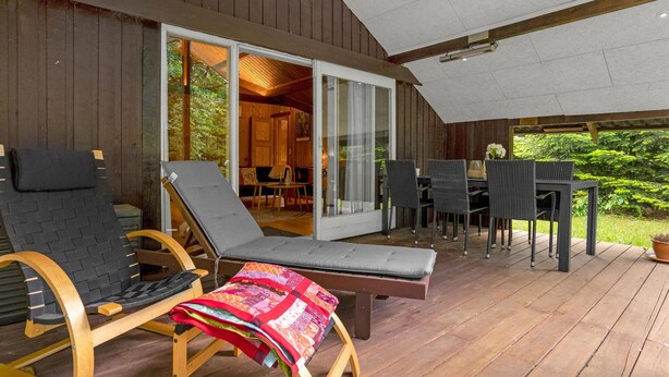 Eine Holzterrasse mit Liegestühlen und einem Tisch, eine Glastür zur Innenraumanbindung und Blick ins Grüne.