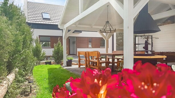 A covered outdoor seating area with a table and chairs, a hanging light fixture, and a view of a house with a sloped roof and wooden shutters. - Amazing home in Nowe Warpno with sauna (Nowe Warpno)