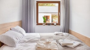 A neatly made bed with white linens, a bedside table with a plant and a speaker, and a window with curtains. - Amazing home in Nowe Warpno with sauna (Nowe Warpno)