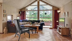 A cozy living room with a high ceiling, wooden walls, a large window, a sofa, a chair, a table, and a television.