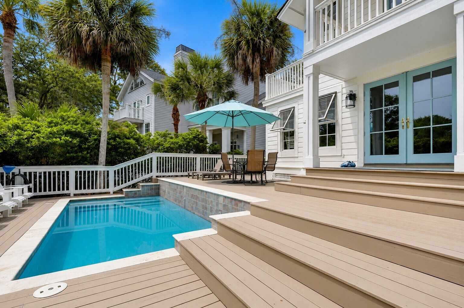 A swimming pool with a wooden deck, a covered seating area with chairs, and a white building with large windows.