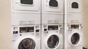 A row of stacked front-loading washing machines with control panels and safety labels. - Travelodge by Wyndham Orange County Airport/ Costa Mesa (Costa Mesa)