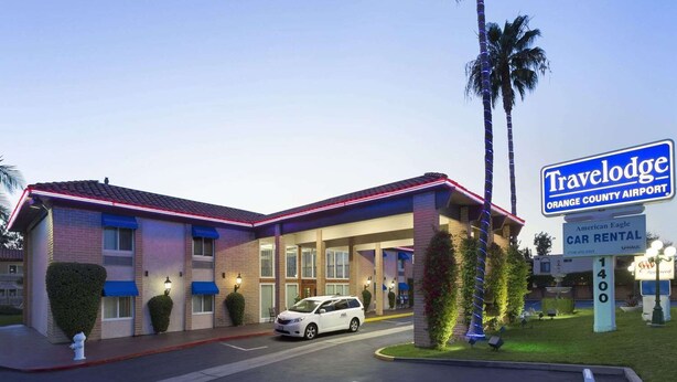 A Travelodge hotel with a car rental sign, a white car parked in front, and a well-lit entrance. - Travelodge by Wyndham Orange County Airport/ Costa Mesa (Costa Mesa)