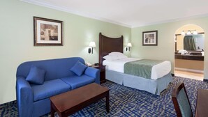 A hotel room with a bed, a blue sofa, a wooden nightstand, a desk, and a bathroom visible through an archway. - Travelodge by Wyndham Orange County Airport/ Costa Mesa (Costa Mesa)