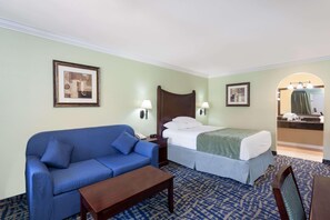 A hotel room with a bed, a blue sofa, a wooden nightstand, a desk, and a bathroom visible through an archway. - Travelodge by Wyndham Orange County Airport/ Costa Mesa (Costa Mesa)