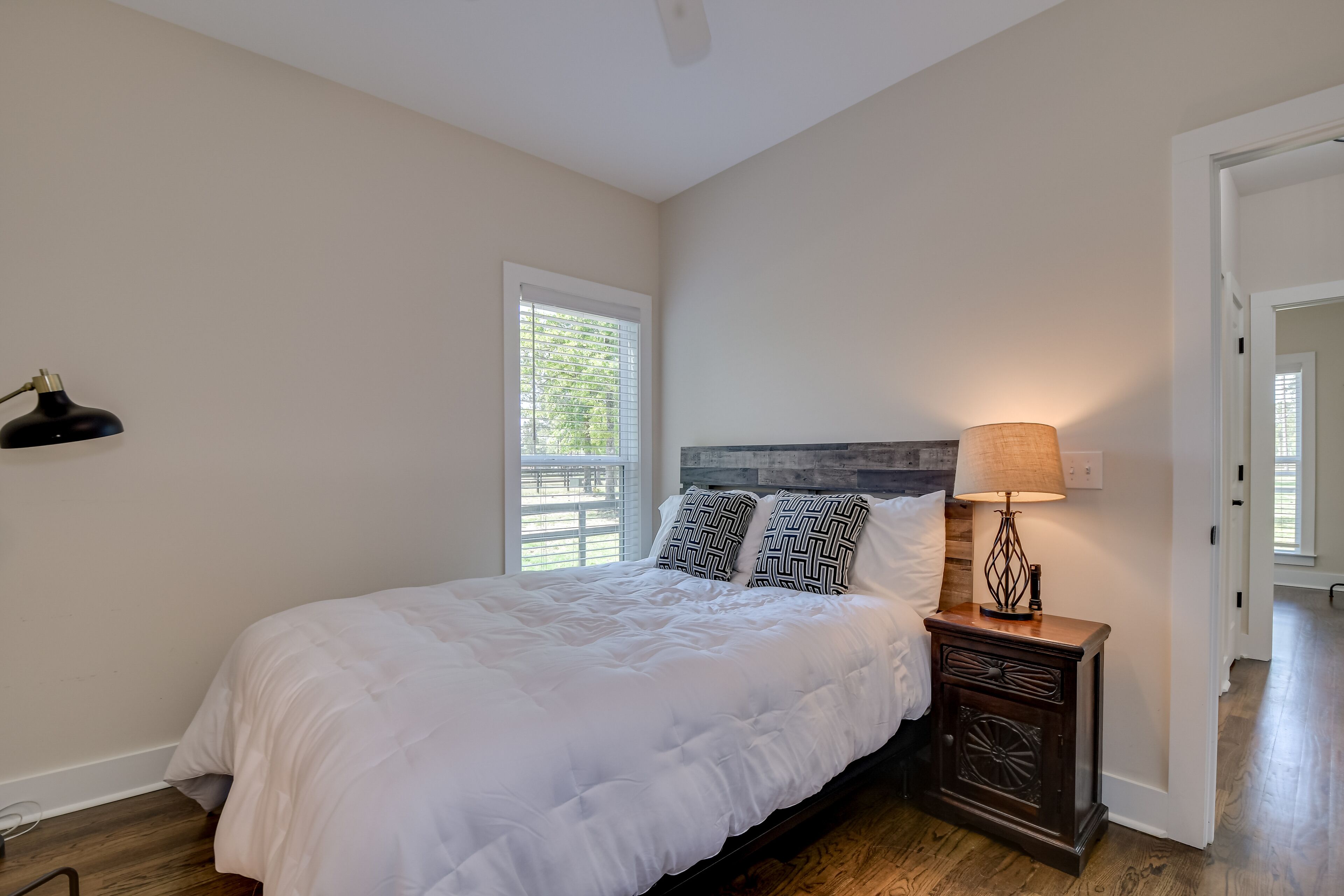 A bedroom with a large bed, a nightstand with a lamp, and a window with blinds.