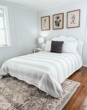 A neatly arranged bedroom with a bed, a nightstand, a lamp, and three framed botanical prints on the wall.