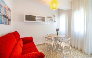 Uma sala de estar moderna com um sofá vermelho, uma mesa de madeira redonda com cadeiras brancas, uma pequena estante com objetos decorativos e uma luminária pendente amarela.