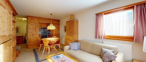 A cozy living room with a sofa, a coffee table, and a dining area with a table and chairs. There is a kitchen area with wooden cabinets and a window with curtains.