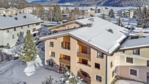 Ein schneebedecktes Gebäude mit Balkon, umgeben von anderen Gebäuden und einer Berglandschaft.