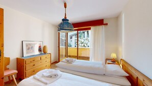 Ein Hotelzimmer mit zwei Betten, einem Holzkleiderschrank, einem gerahmten Bild und einem Fenster mit Blick auf Bäume.