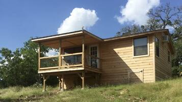 A wooden cabin with a balcony and a second-story deck, surrounded by trees and grass.