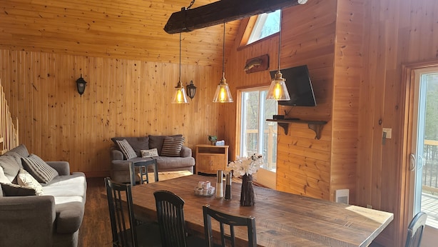 A cozy cabin interior with a wooden interior, a sofa, a dining table, and a TV mounted on the wall. - Waterfront Family Cottage! (Notre-Dame-de-Pontmain)