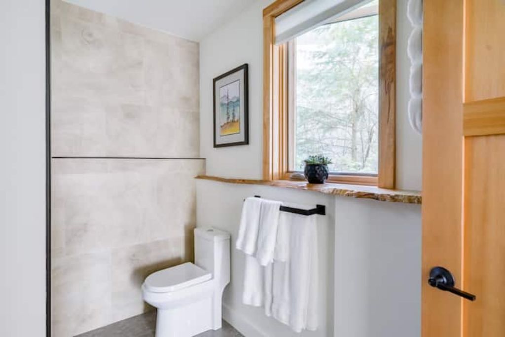 A bathroom with a toilet, a towel rack with white towels, a framed picture on the wall, and a window with a view of trees.