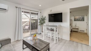 A modern living room with a flat-screen TV, a dining area with white chairs, and a ceiling fan.