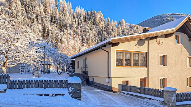 A two-story chalet with a stone staircase leading to the entrance, surrounded by snow-covered trees and a clear blue sky.