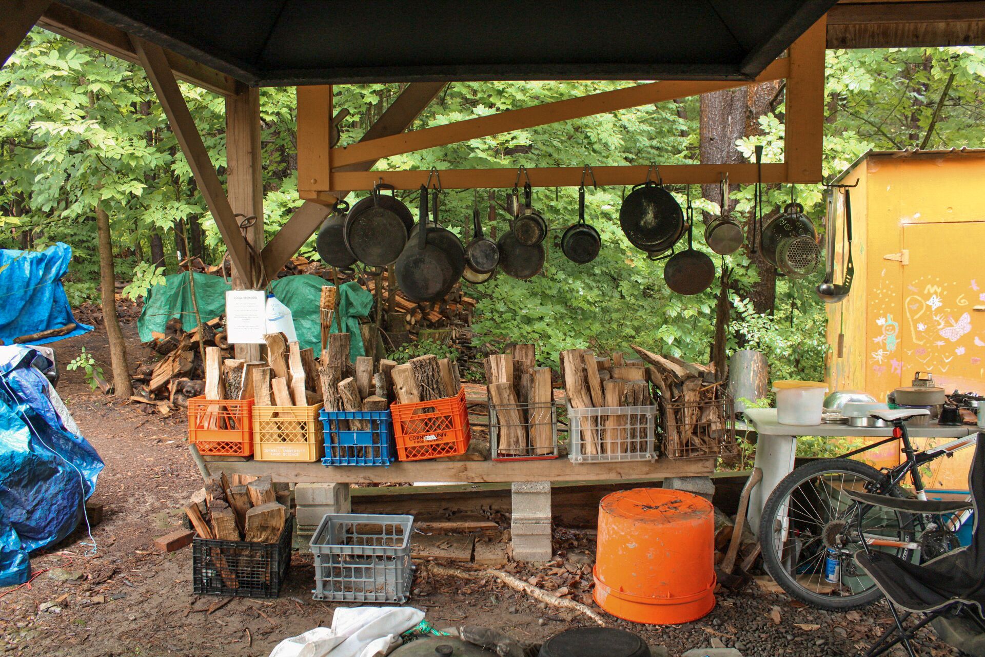 Cucina esterna rustica con cassette di legna, pentole appese e una bicicletta.