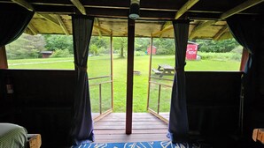 A room with a wooden ceiling, a bed, a blue patterned rug, and a view of a grassy area with a picnic table and a red building in the distance.