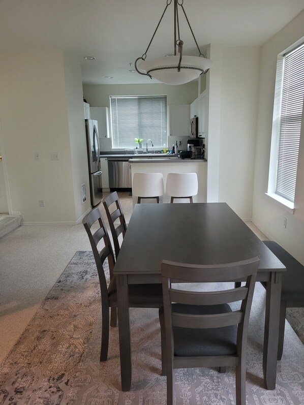 Un comedor con una mesa y sillas de madera oscura, una alfombra estampada y vistas a la cocina con una ventana y persianas.