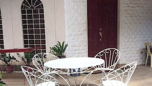 Une table en métal blanc pour l’extérieur, accompagnée de quatre chaises, devant un mur en briques blanches et une porte rouge en arrière-plan.