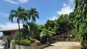 Un quartier résidentiel avec une allée pavée, une voiture garée et une végétation luxuriante comprenant des palmiers et des plantes à fleurs.