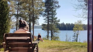Eine Person sitzt auf einem hölzernen Stuhl und blickt auf einen See mit Bäumen im Hintergrund.