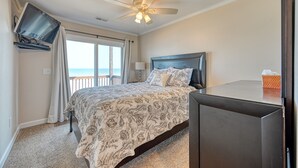 Quarto com cama, ventoinha no teto, TV fixada na parede e vista para o oceano através de uma janela grande.