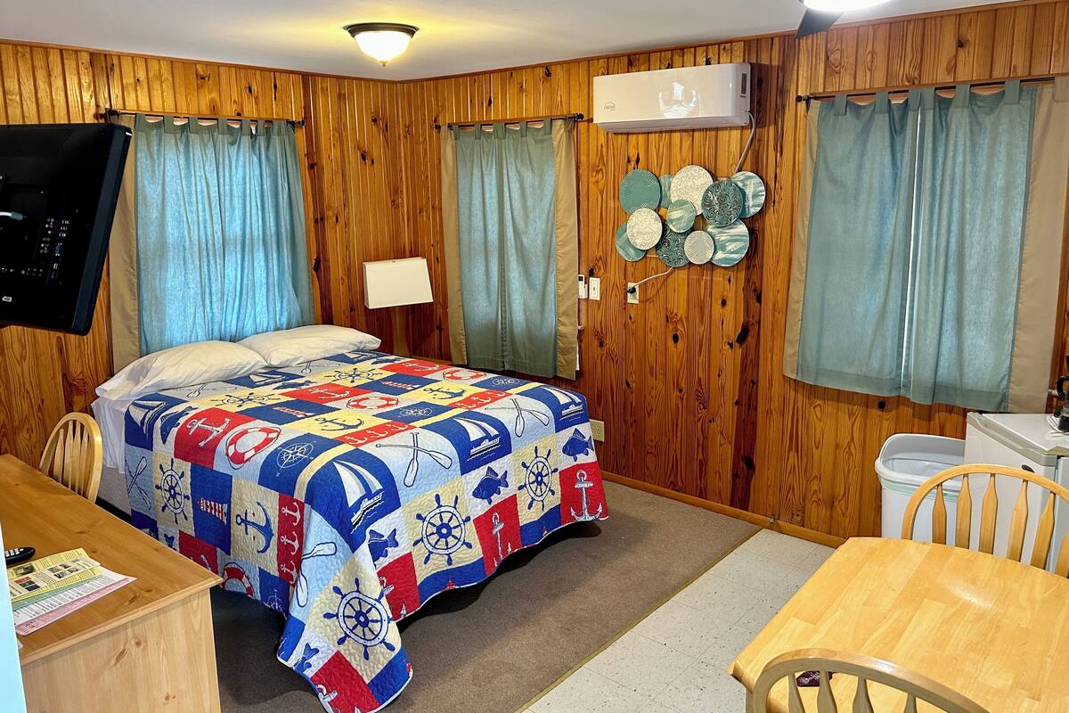 Una habitación con pared de paneles de madera, una cama con colcha de temática náutica, un escritorio con un control remoto, una silla, un aire acondicionado de pared y un ventilador de techo.