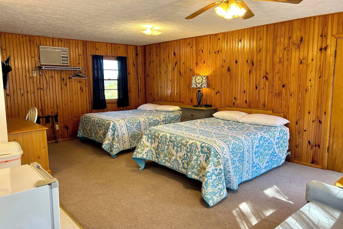 Una habitación con dos camas, ventilador de techo, calefactor, ventana con cortinas y una lámpara en una mesita de noche.