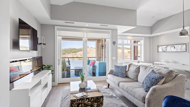 A modern living room with a large sofa, a coffee table, and a television. There is a sliding glass door leading to a balcony with outdoor furniture.