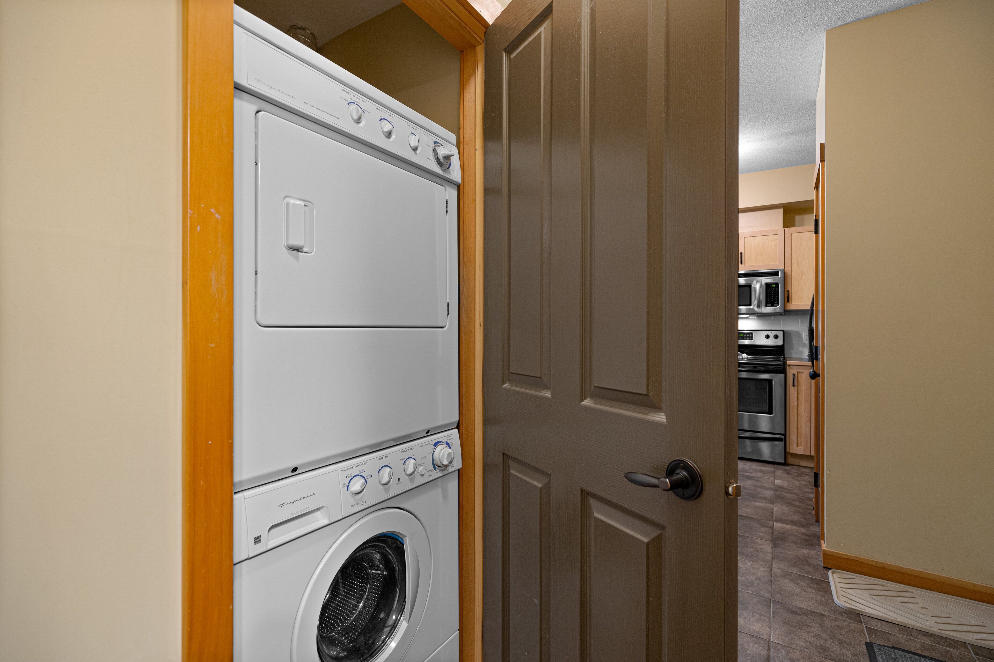 Une buanderie avec une machine à laver et un sèche-linge superposés, une porte en bois et, au fond, une cuisine.