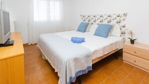 A bedroom with a bed, a TV on a wooden desk, a nightstand with a plant, and a window with curtains. - Beautiful apartment for 4 guests with WIFI, pool, TV and balcony (Santa Susanna)