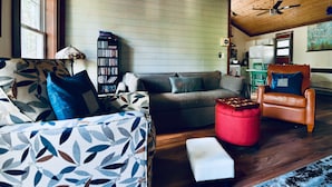 Una sala de estar acogedora con sofá, reposapiés y sillón con cojín rojo. Se observa una estantería contra la pared y un ventilador de techo.