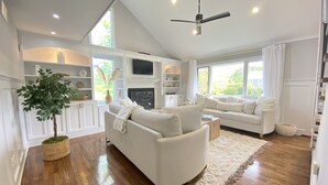 A spacious living room with a white sofa, a wooden floor, a ceiling fan, and a large window providing natural light. - 5BR, Luxury Home, Pool, Large Yard , Close to Beach & Town (Southampton)