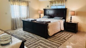 A bedroom with a dark wooden bed, two bedside tables with lamps, a large window with curtains, and a ceiling fan. - Tropical Getaway Home (Frederiksted)