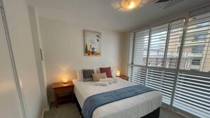 Une chambre à coucher comprenant un lit, deux tables de chevet, une commode, une fenêtre avec des stores et un luminaire au plafond.