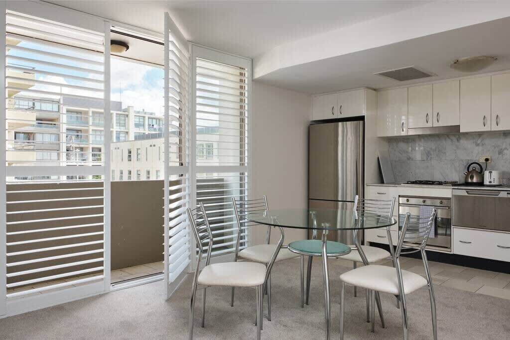 A modern kitchen with white cabinets, a stainless steel refrigerator, and a glass dining table with four chairs.