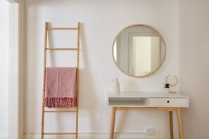A white desk with a round mirror, a small plant, and a white pitcher. A wooden ladder with a pink towel is leaning against the wall.
