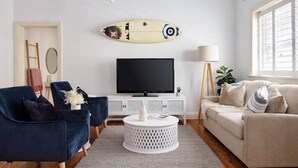 Un salon comprenant un téléviseur, deux fauteuils, un canapé, une table basse ronde et une décoration murale représentant une planche de surf.