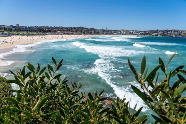 A coastal beach scene with waves, a clear sky, and buildings in the background.
