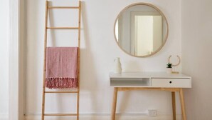 Un bureau blanc orné d’un miroir rond, d’une petite plante et d’un pichet blanc. Une échelle en bois, sur laquelle est posée une serviette rose, est appuyée contre le mur.