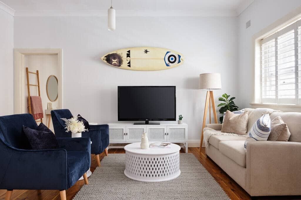 A living room with a TV, two chairs, a sofa, a round coffee table, and a surfboard decoration on the wall.